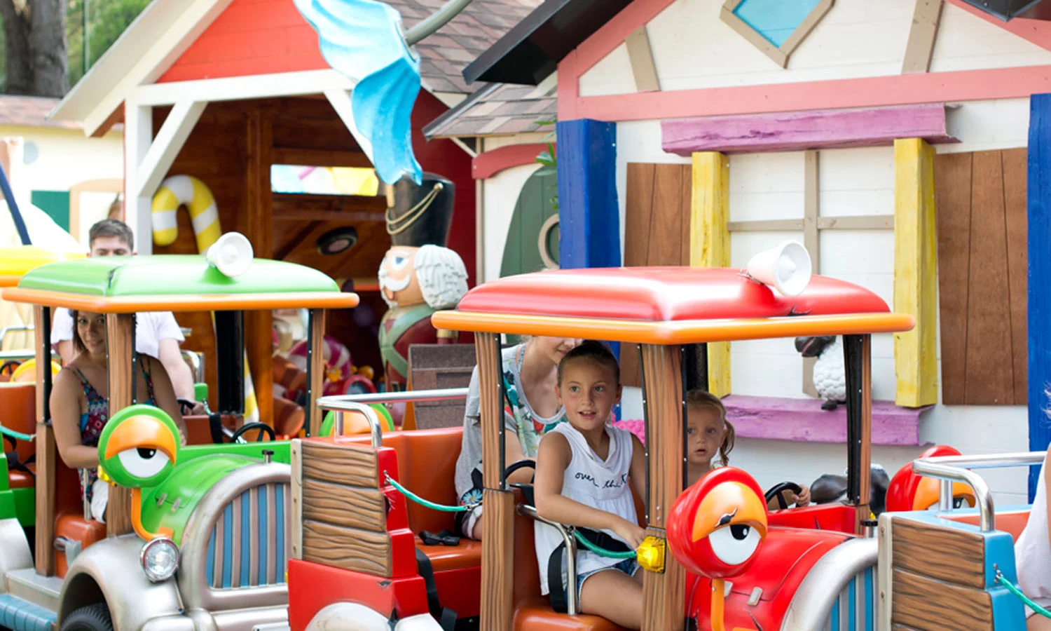 Colorful mini-trucks carrying children, driving through a whimsical outdoor amusement park near small houses and animal statues at Union Lido