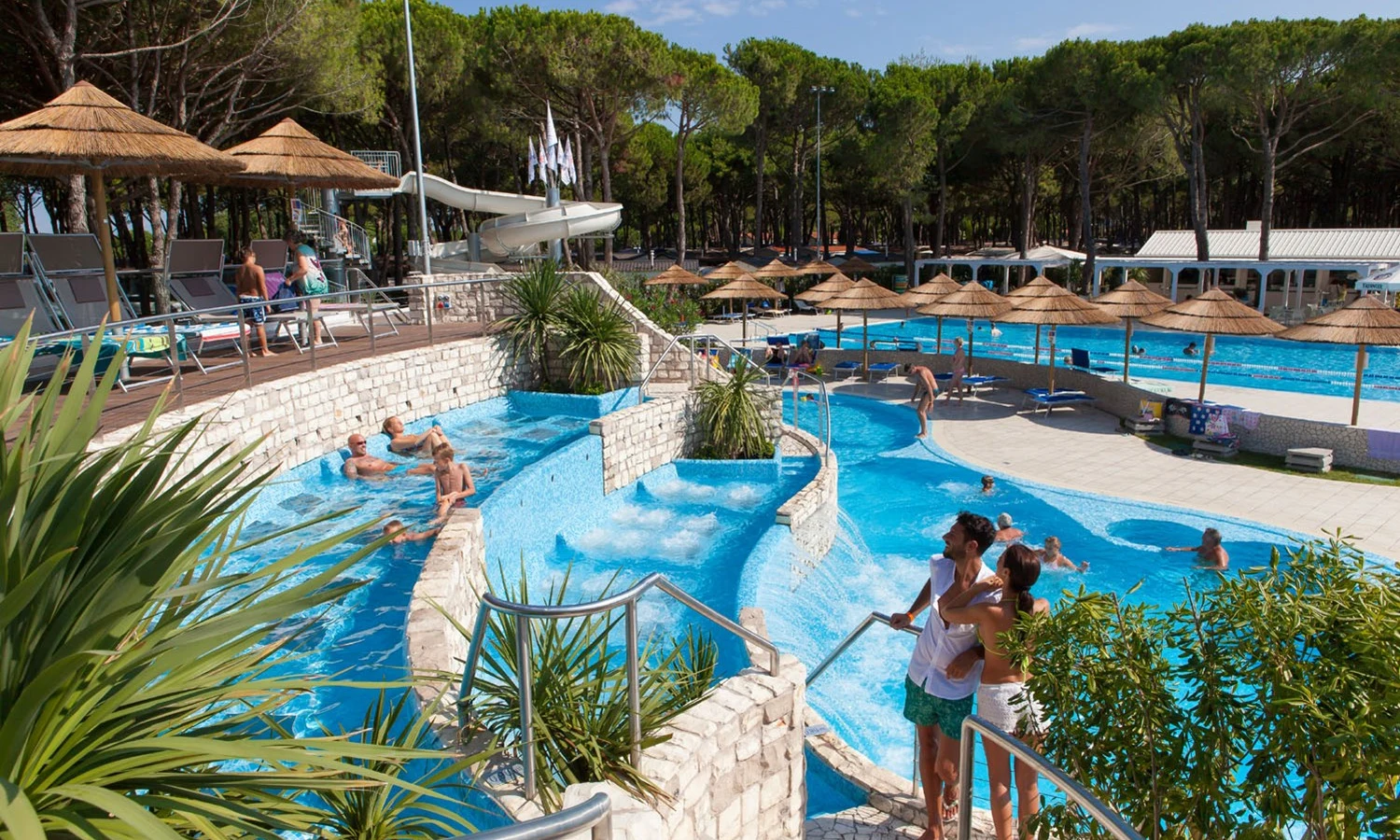 People relaxing and swimming in multi-level pools with slides, surrounded by trees and sun loungers at Ca'Pasquali