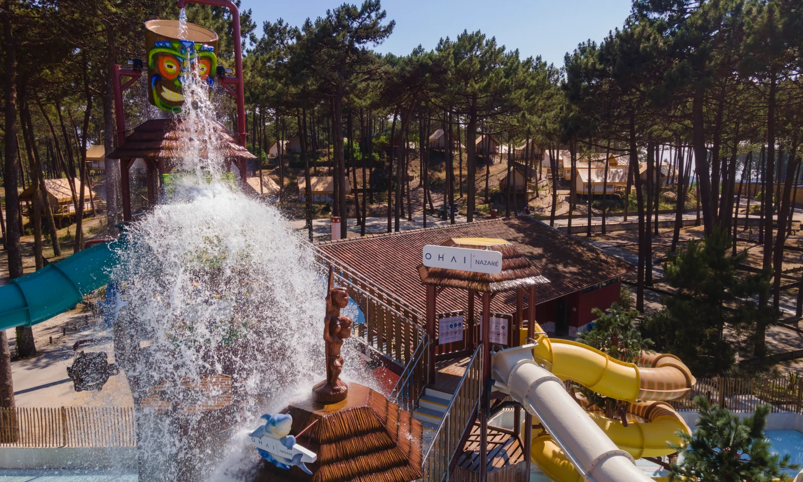Waterpark splash-structure tipping large water bucket amidst tall pine trees, surrounded by slides and rustic cabins at Ohai Nazaré Outdoor Resort