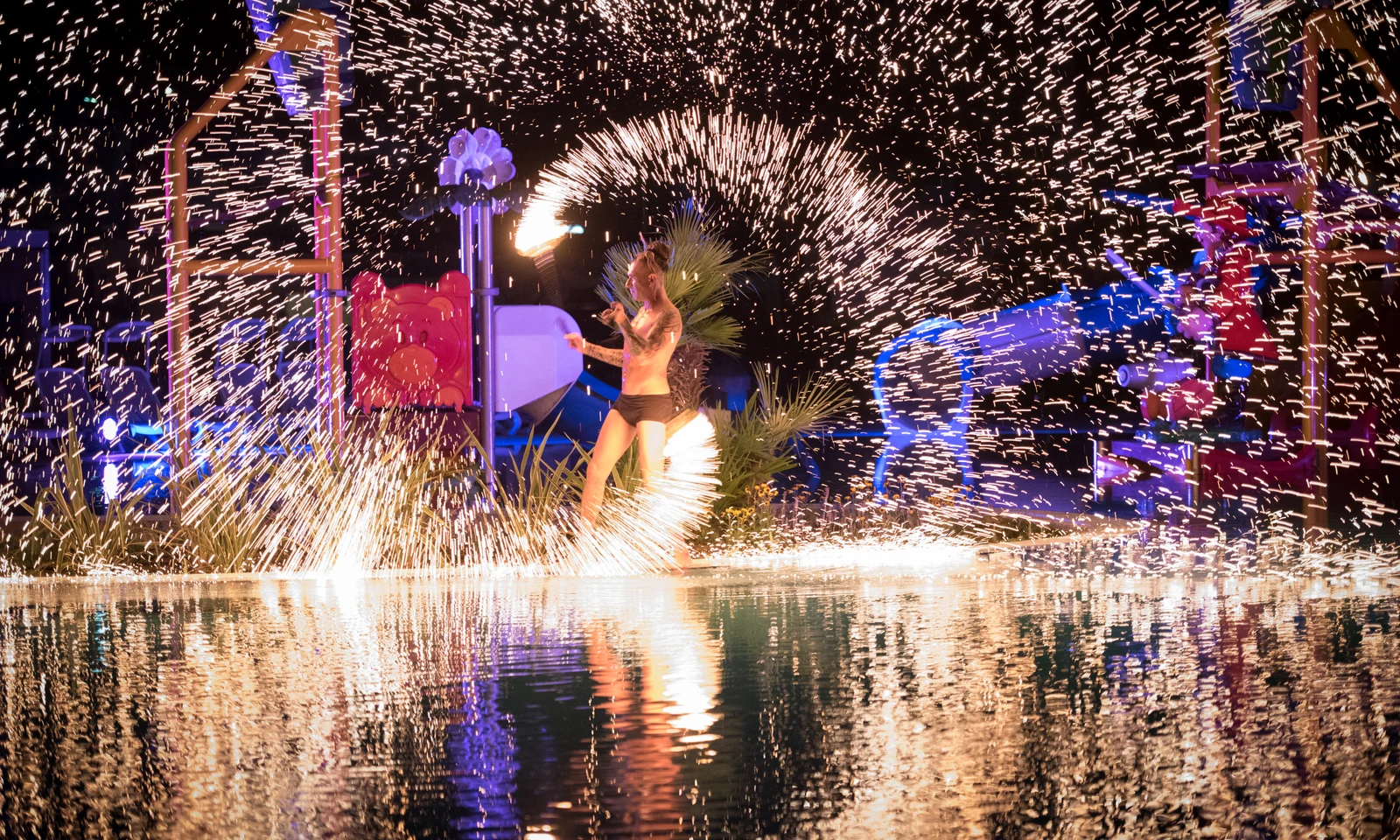 Performer spinning fire baton, surrounded by bright sparks and night-lit playground, reflected in water at Residence Village
