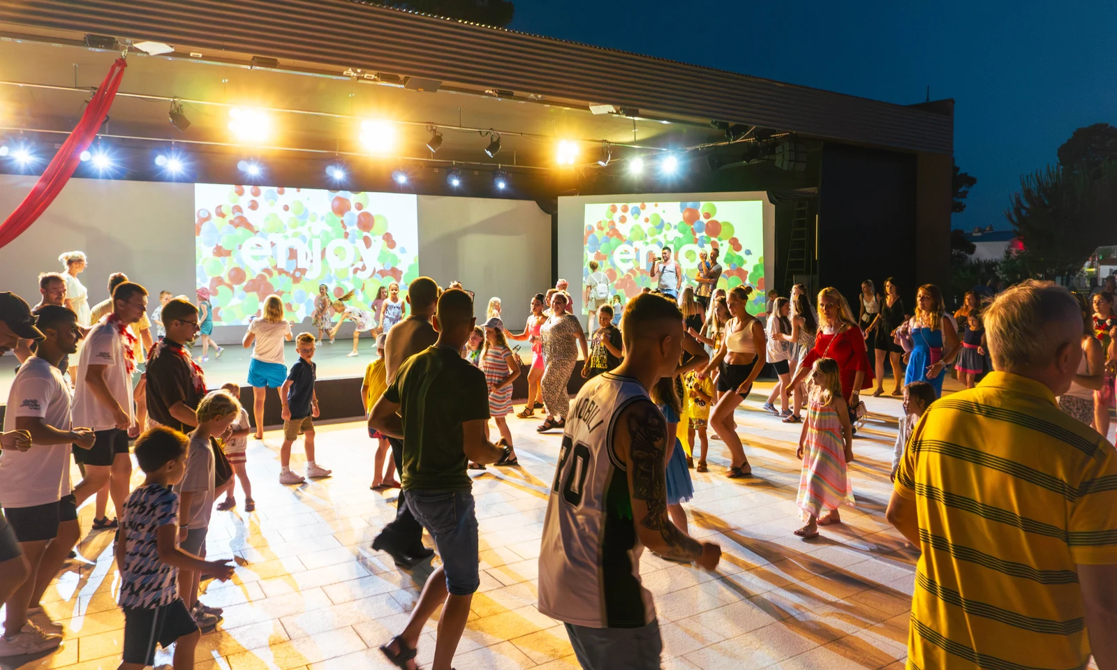 People dancing energetically under bright stage lights during an outdoor evening event, with colorful visuals on a screen at Zaton Holiday Resort