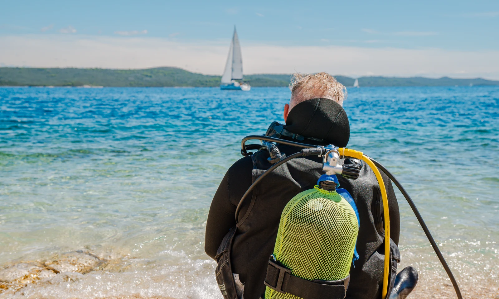 Scuba diver sits by the shore, facing the sea with a sailboat in the distance at Zaton Holiday Resort