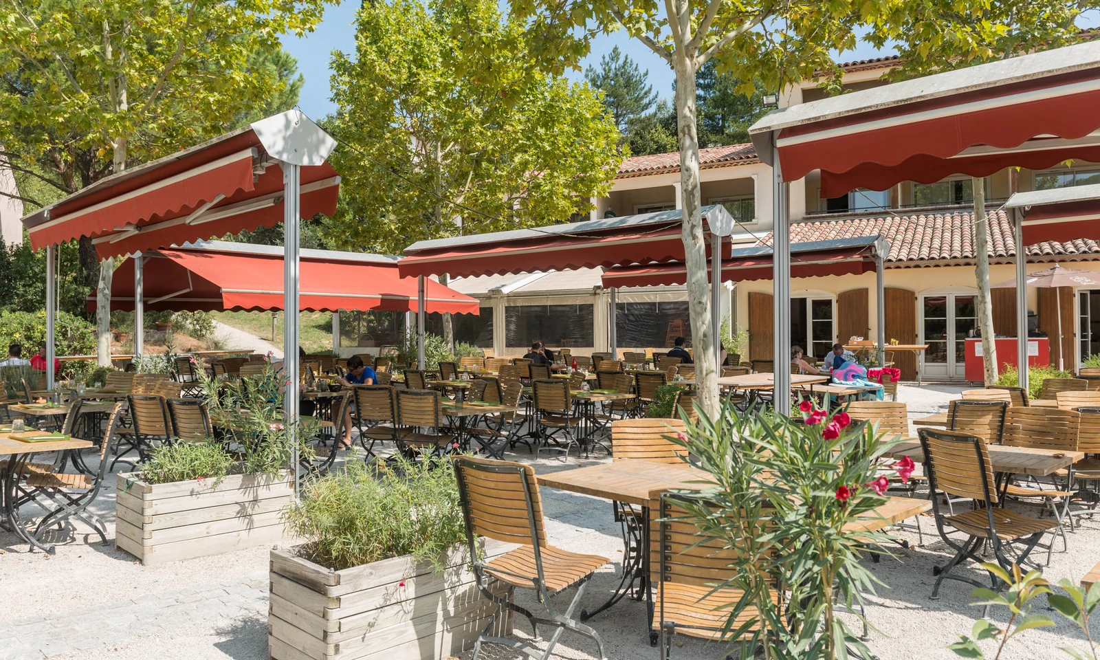 Wooden tables and chairs under red awnings sit empty outside a building with trees and people around at Le Rouret en Ardeche