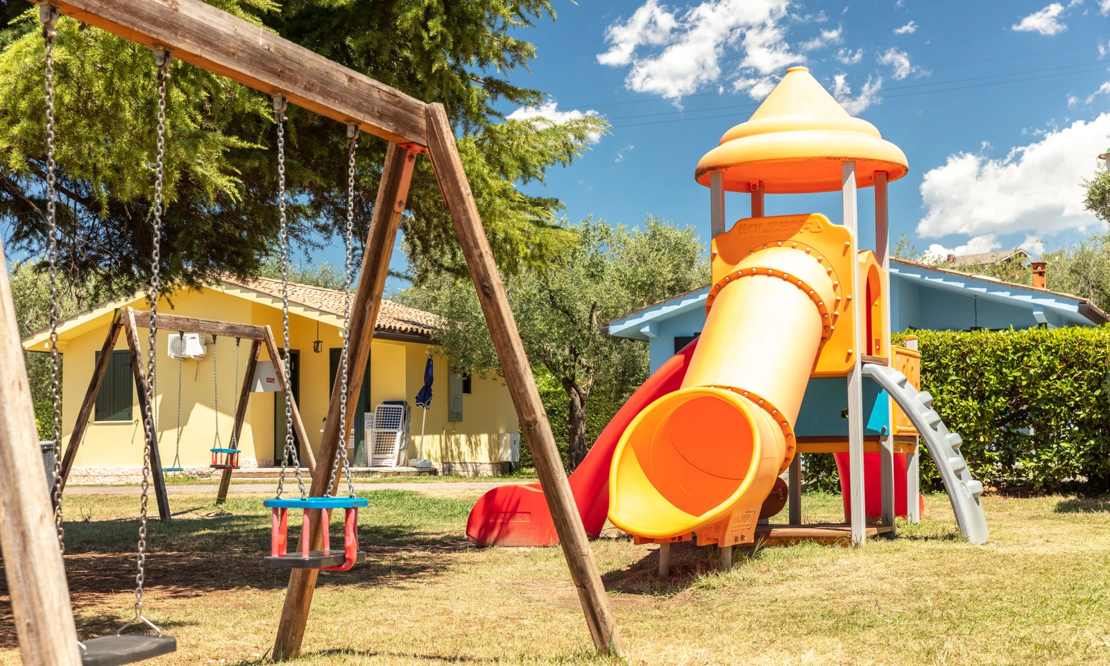 Playground structure with slide and swingset on grass, surrounded by trees and colorful buildings at Cisano/San Vito