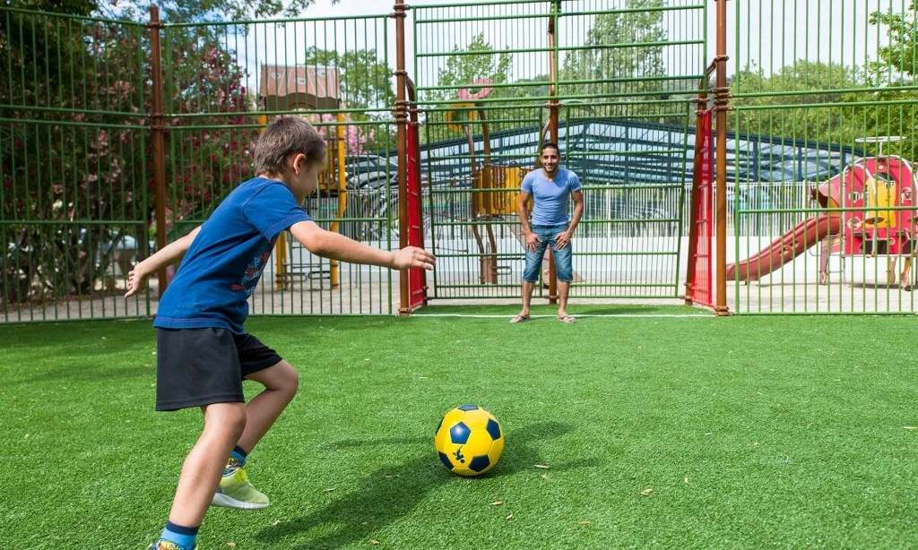 Child kicking a soccer ball towards adult standing as goalkeeper in an outdoor fenced playground at Le Saint Louis