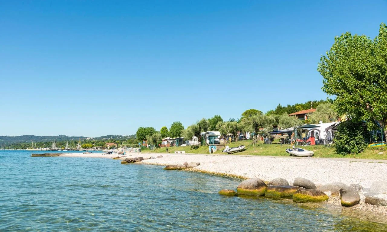 Boats rest on a rocky beach beside a clear lake, lined with trees and tents, under a clear blue sky at Fontanelle