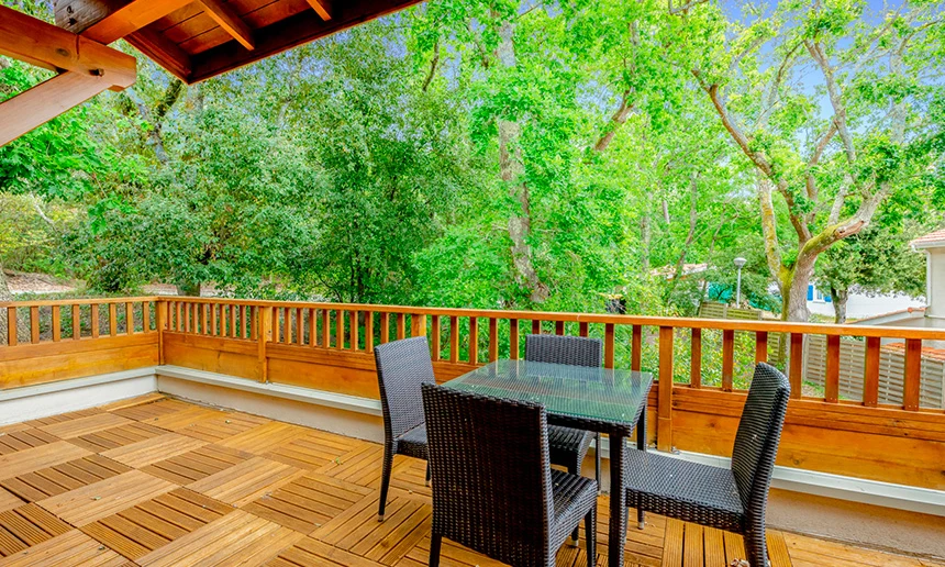 Four wicker chairs surround a glass table on a wooden deck with a view of lush, green trees at Residence Les Rives du Lac