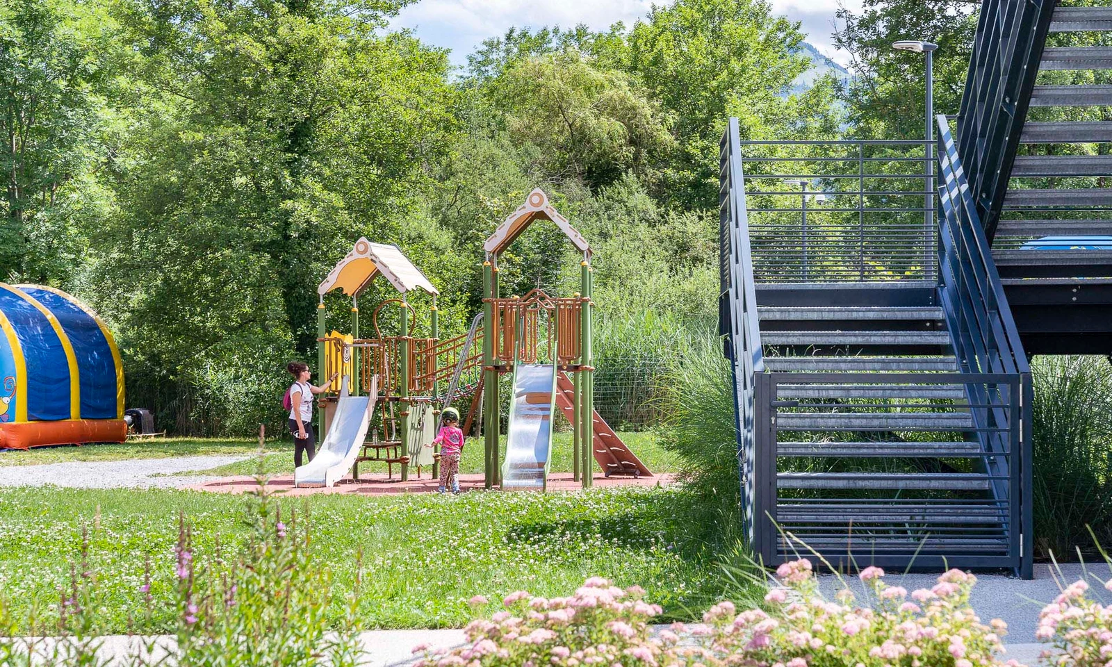 Children playing on slides and climbing equipment, next to an outdoor staircase, in a lush green park at La Nublière