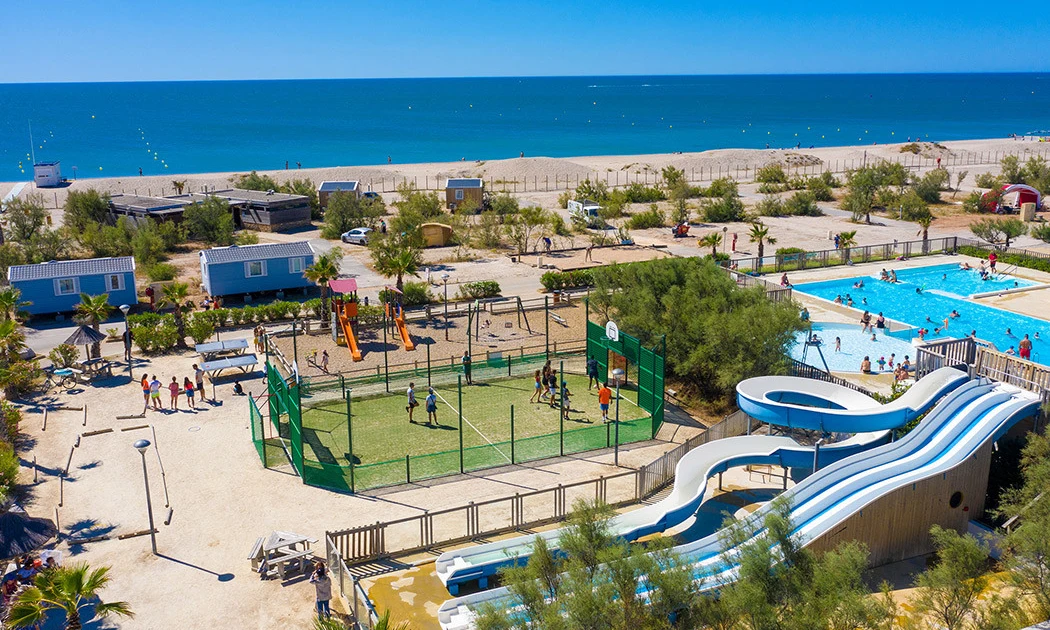 Swimming pools, slides, and sports court amidst trees, people, and mobile homes near the beach at Marvilla Parks Le Palavas