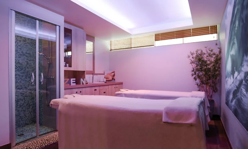 Two massage tables with white linens are situated in a dimly lit treatment room, featuring a shower and bamboo decor at Houlgate Premium Residence et Spa