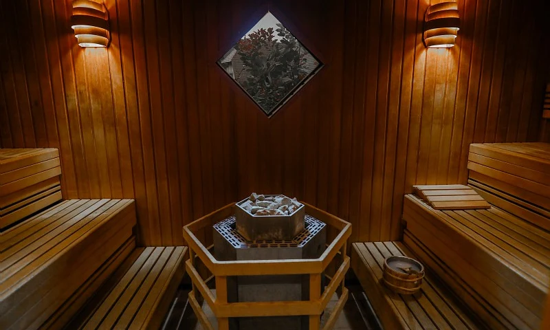 Wooden benches surround a central heater with stones in a well-lit sauna room, featuring a small window at Houlgate Premium Residence et Spa
