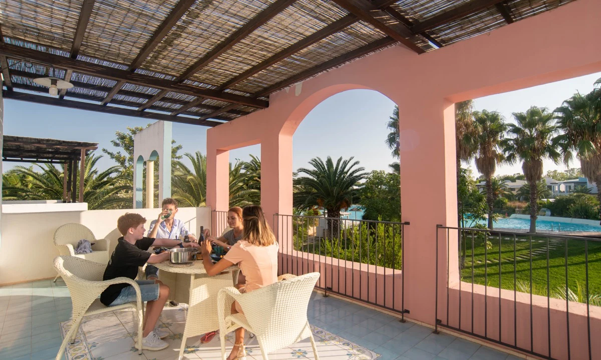 People chatting and eating around a table on a covered terrace, surrounded by a garden with palm trees and a pool at I Turchesi Club Village