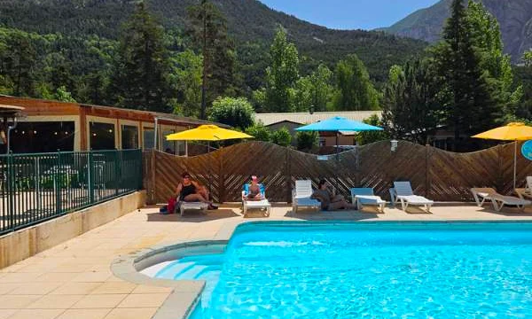 Pool with lounge chairs under umbrellas by a wooden fence, nestled in a mountainous, forested campsite at Paradis Les Relarguiers
