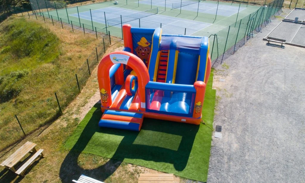 Large inflatable bounce house with slide, situated near tennis courts and picnic benches, surrounded by grass and gravel paths at Caravanile