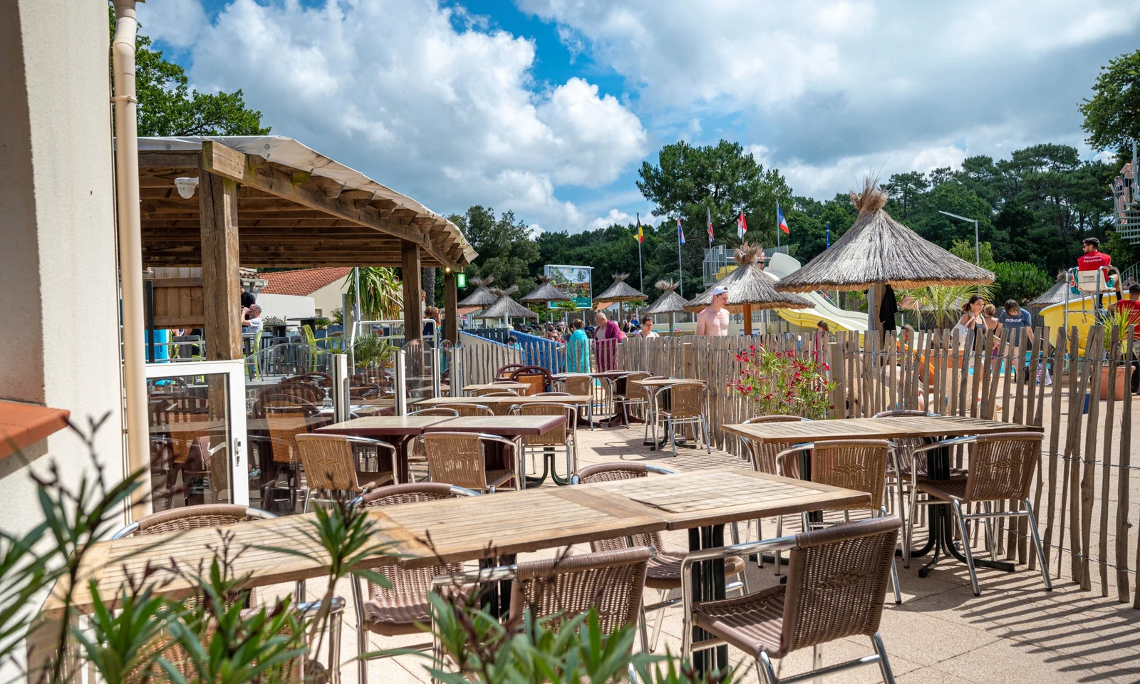 Wooden tables and wicker chairs under a canopy, near a pool area with thatched umbrellas, people, and flags at Flower Camping Les Biches