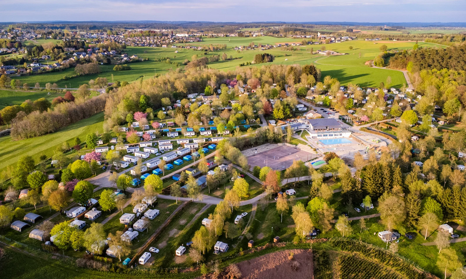 Campers and RVs parked among trees, surrounding a pool complex, with a village and green fields in the distance at Ardennen Camping Bertrix