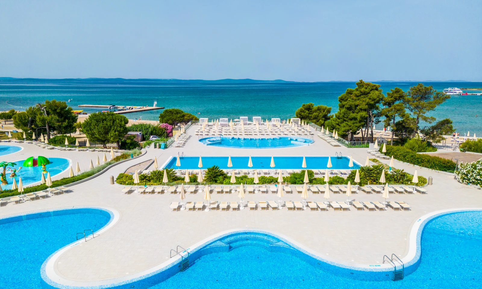 Loungers and parasols surround multiple swimming pools beside the turquoise sea, bordered by lush greenery at Zaton Holiday Resort