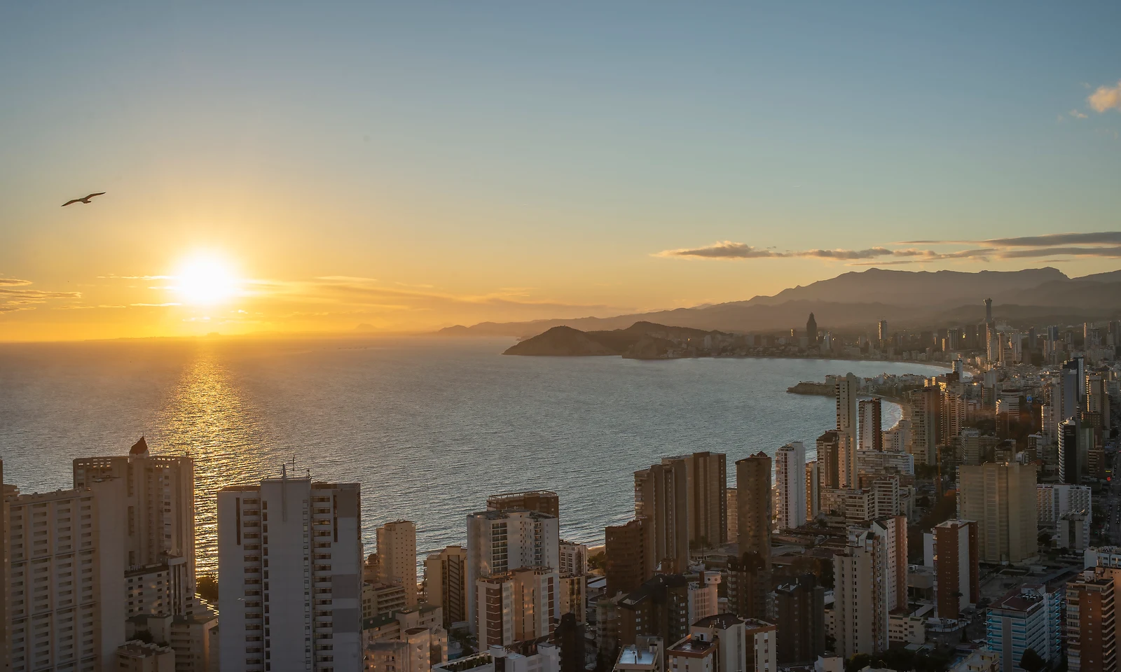 The sun setting over the sea adjacent to a cityscape of tall buildings and distant mountains at Benidorm Horizon