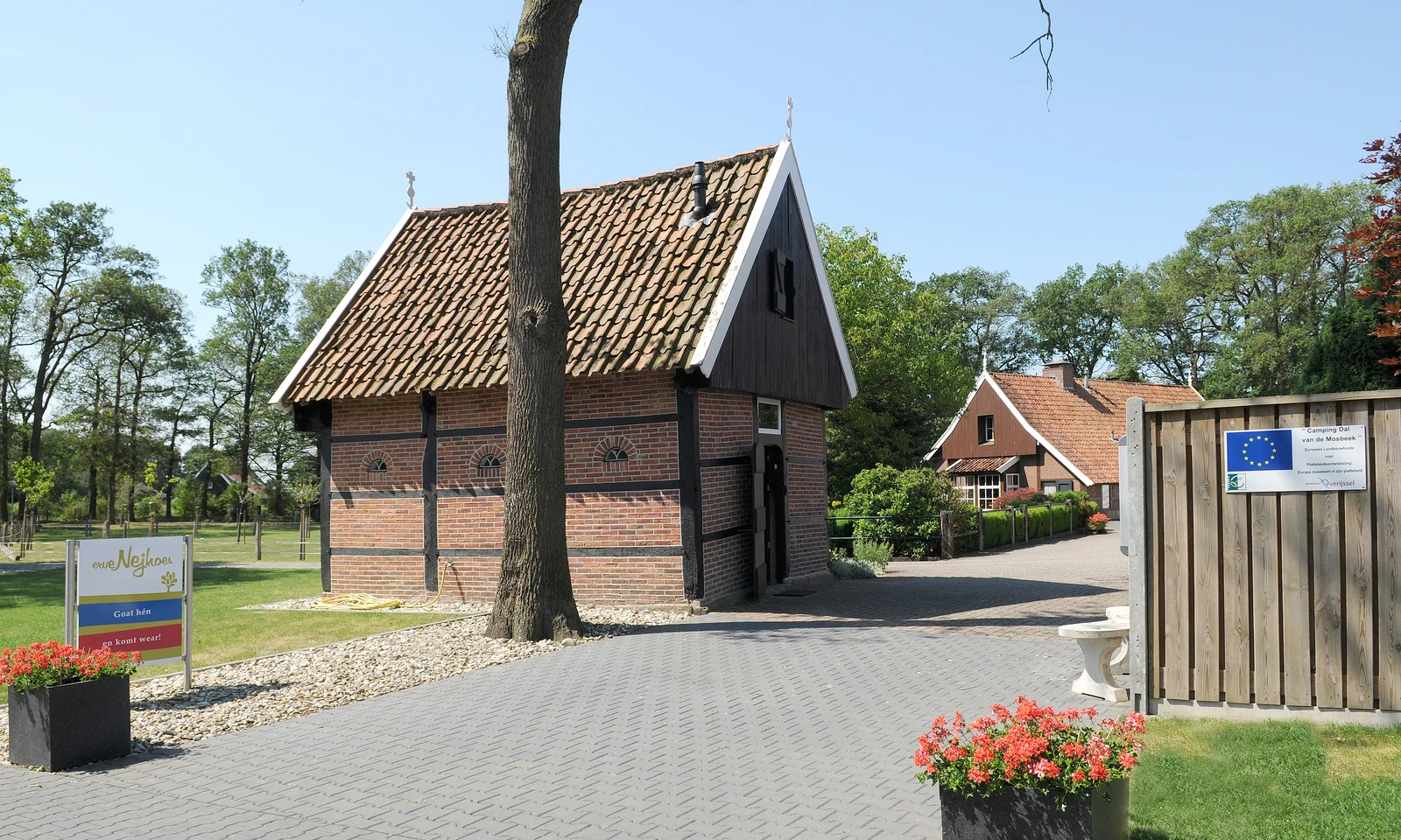 Brick building with tiled roof surrounded by trees and pathway, two potted plants with flowers in foreground at Dal van de Mosbeek