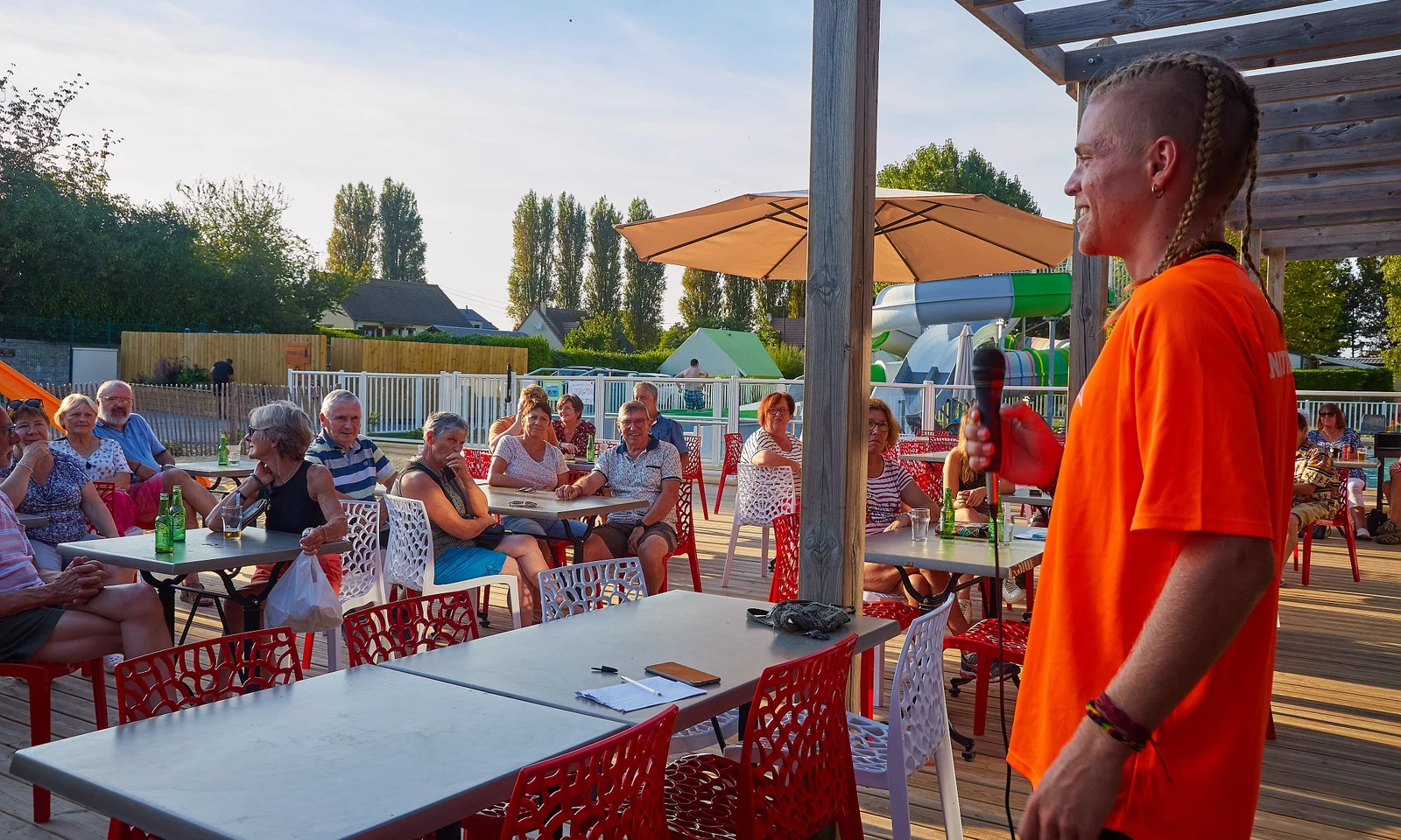 Person speaking into a microphone to an audience seated at outdoor tables under umbrellas at Marvilla Parks Riva Bella