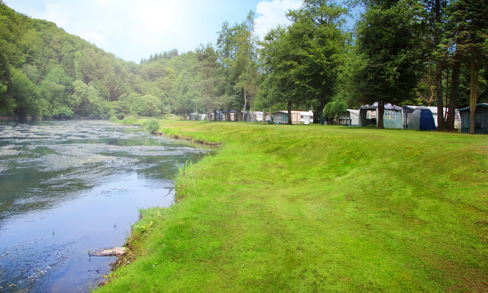 River flows beside grassy bank, with tents and trailers nestled among trees at d'Houlifontaine