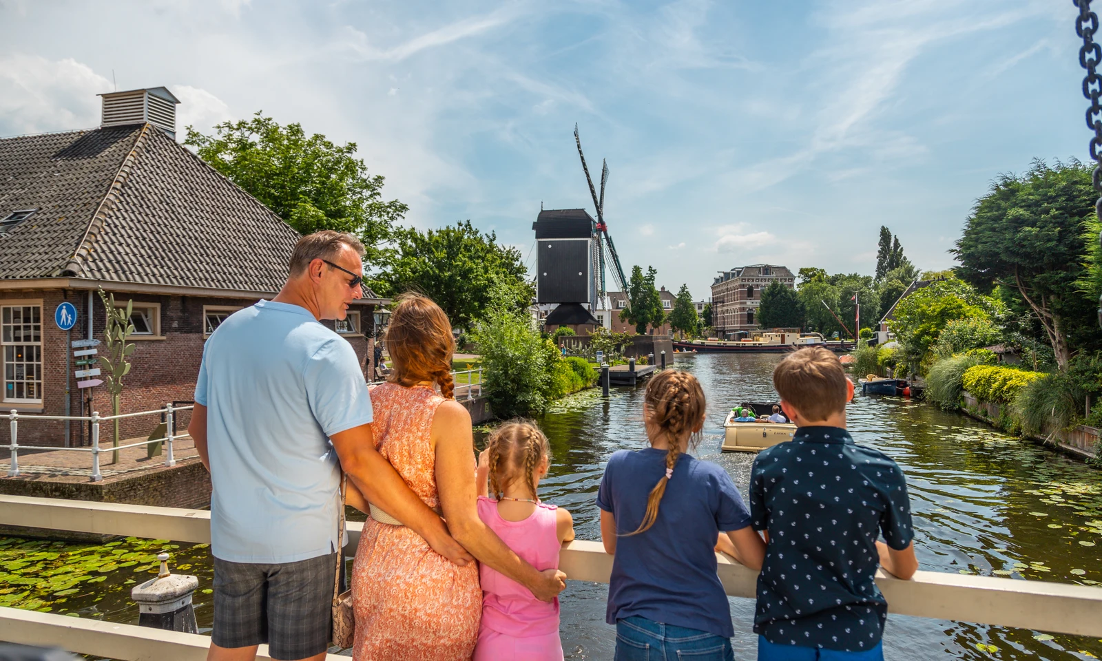 Family watching a boat on a scenic canal, surrounded by greenery and buildings, including a windmill at Europarcs Parc du Soleil