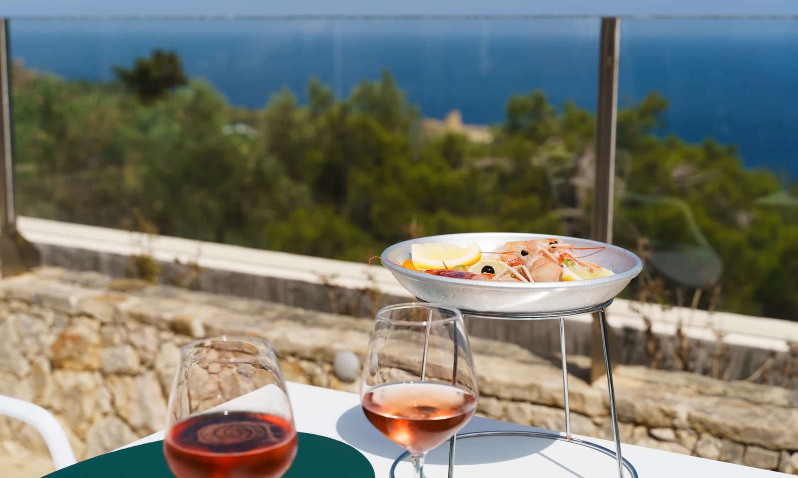 Seafood platter on a stand beside two glasses of rosé, overlooking a stone wall and ocean view at La Scogliera