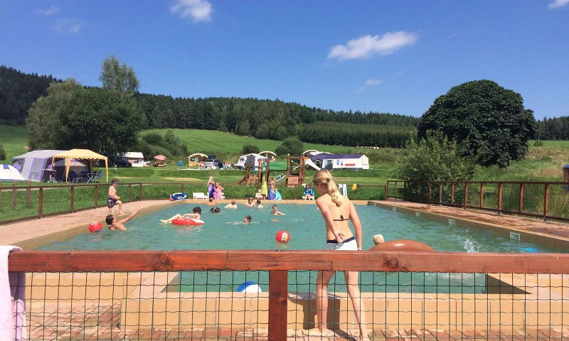 Children swimming and playing with water toys in a pool surrounded by campers and lush, green hills at Chvalsiny