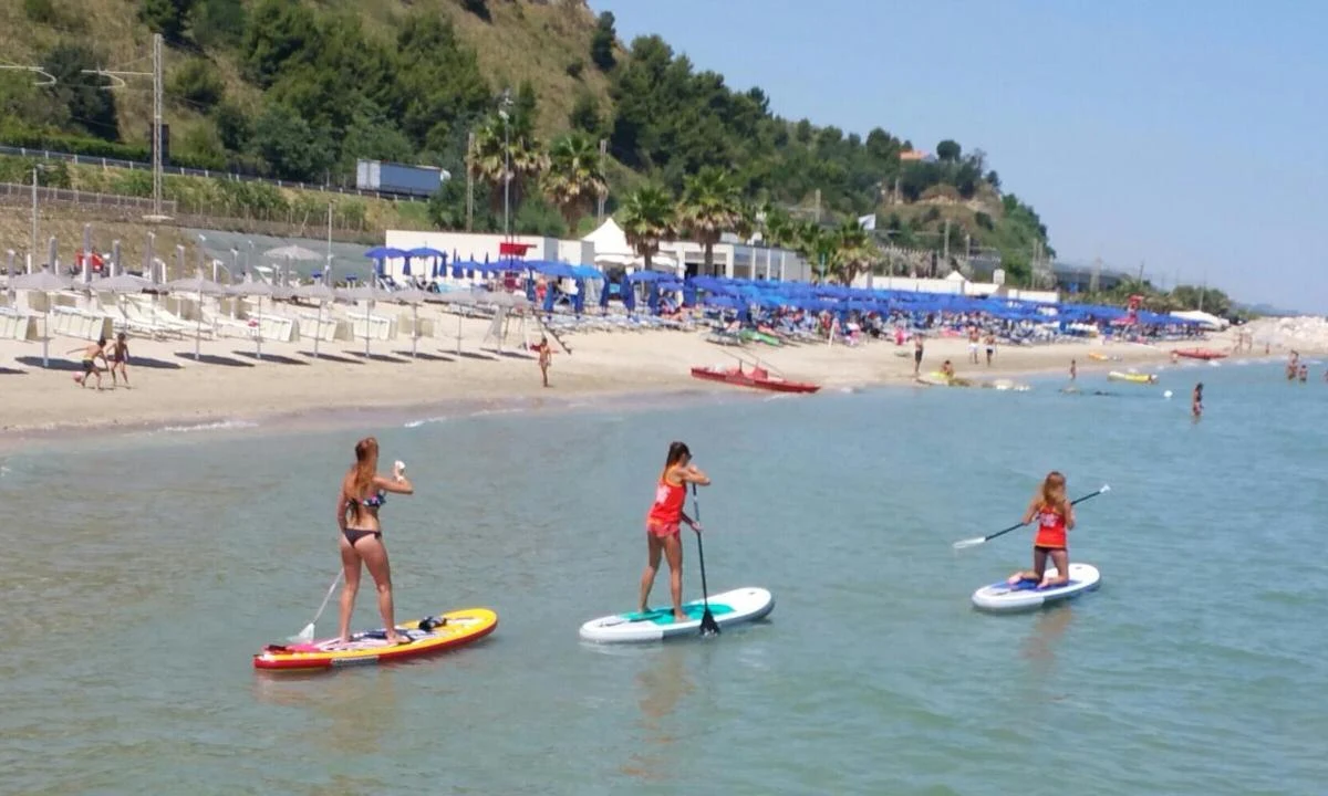 Three people paddleboarding on calm sea near a crowded beach with umbrellas and distant greenery at Camping Village Mirage