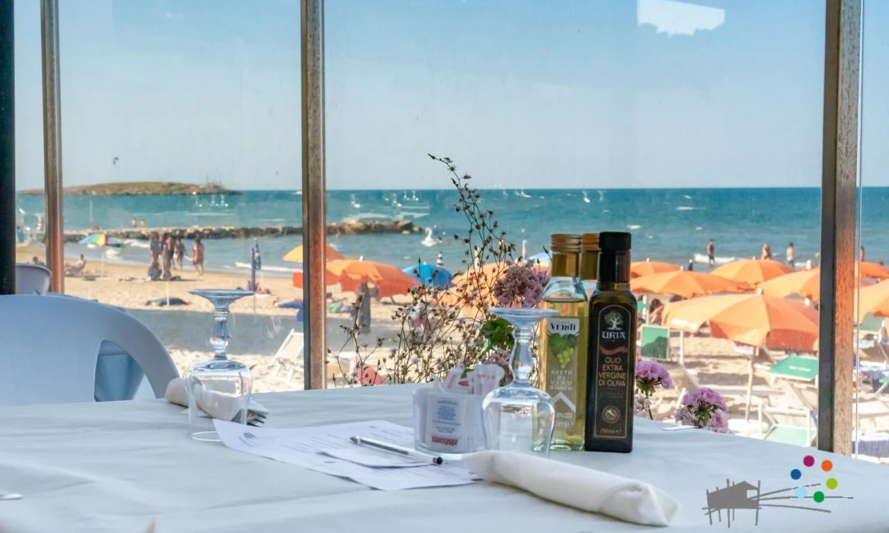 Laid table with olive oil and wine glasses, beach with orange umbrellas and people in background at Spiaggia Lunga