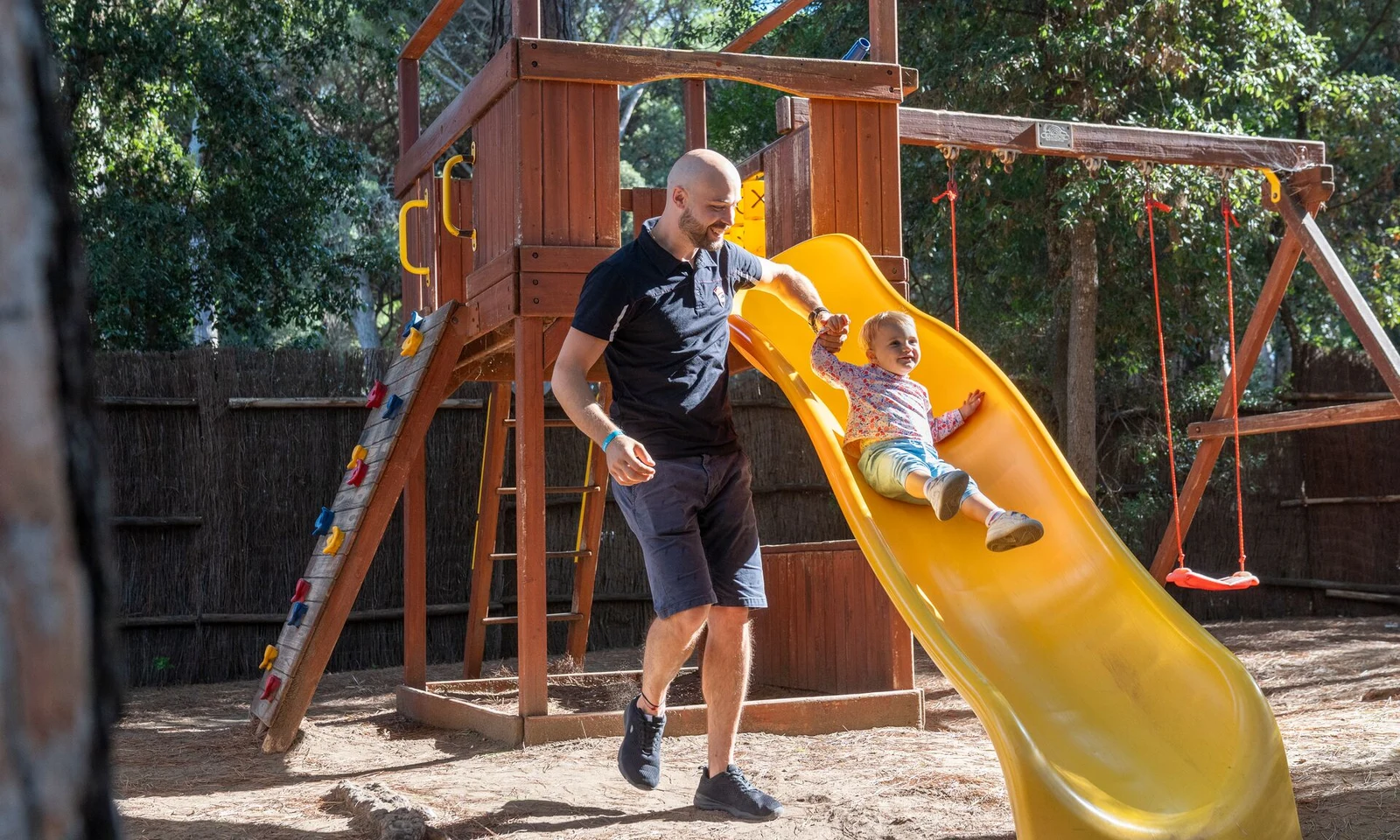 A child slides down a yellow slide while an adult assists in a wooded playground at Etruria Camping Village