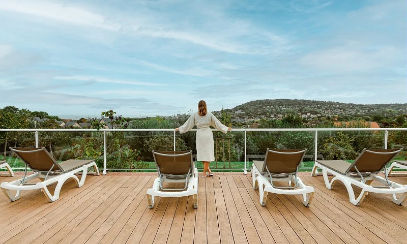 Person standing on a wooden deck with lounge chairs, overlooking a scenic landscape with hills and greenery at Houlgate Premium Residence et Spa