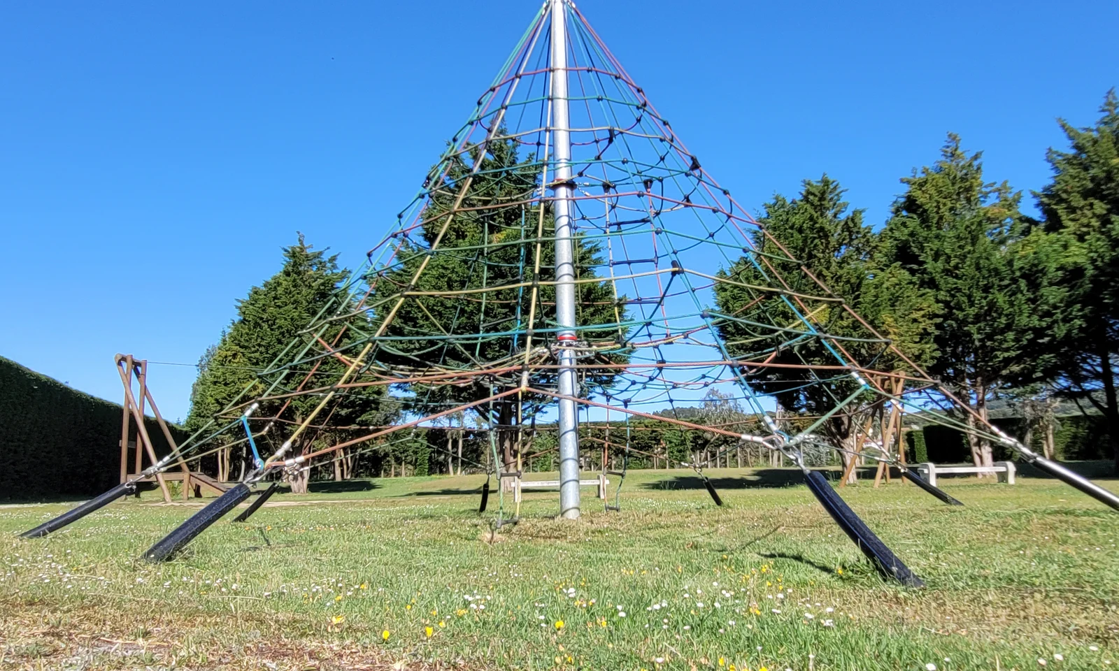 Pyramid-shaped climbing net structure stands on grass with trees in the background at Playa Joyel