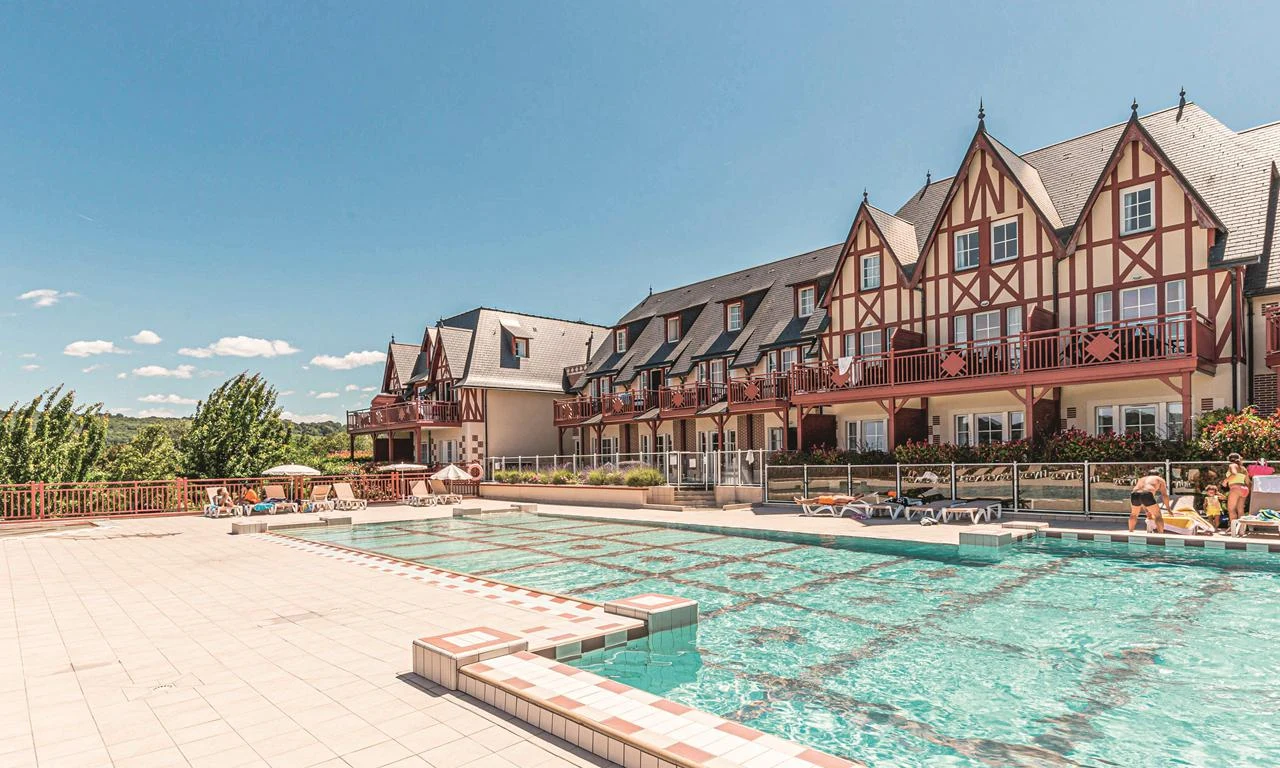 A large outdoor swimming pool with people relaxing beside it, adjacent to a quaint, multi-story building at Houlgate Premium Residence et Spa