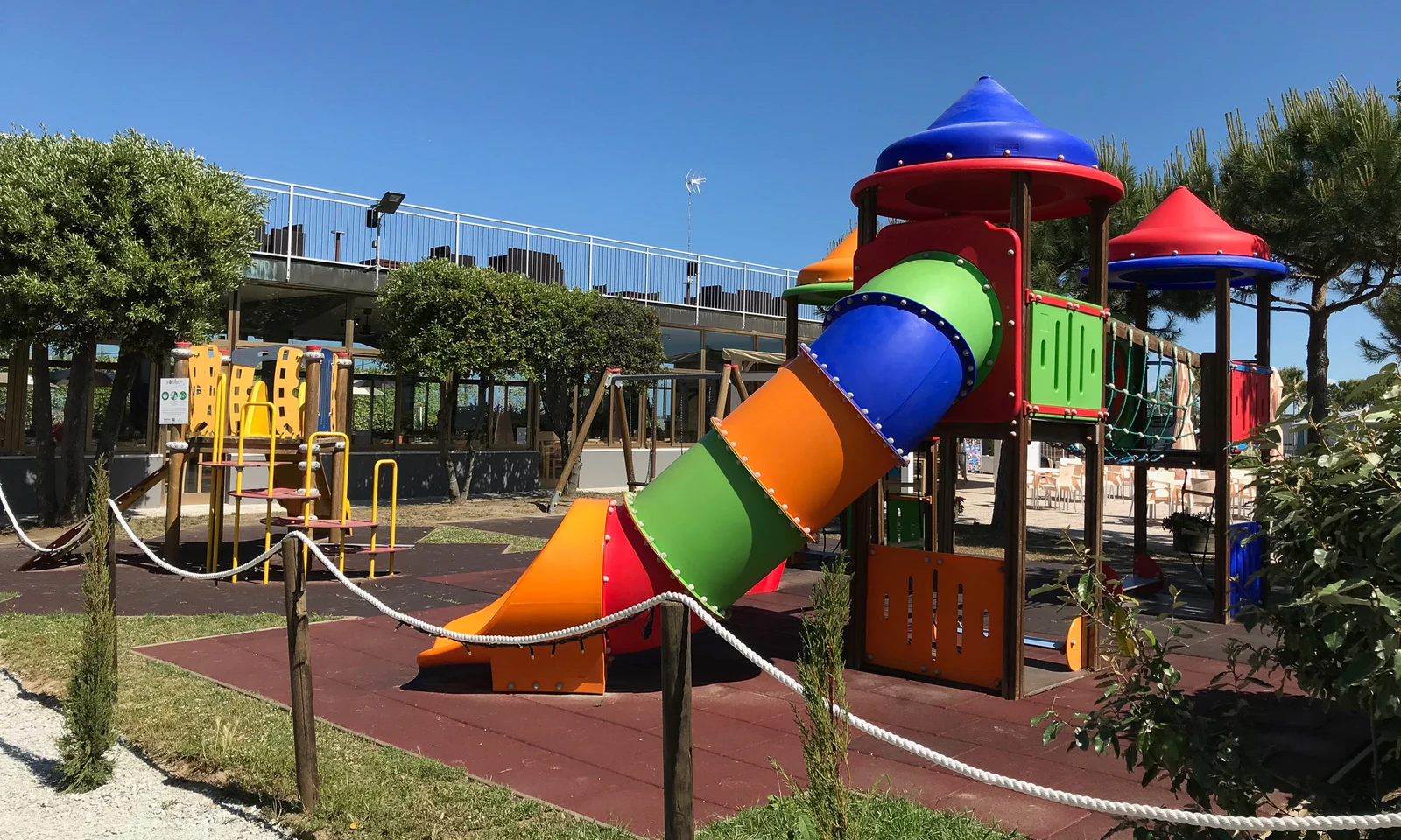 Colorful playground structure with slides and climbing areas, surrounded by green trees and clear blue skies at Pini Beach Village 
