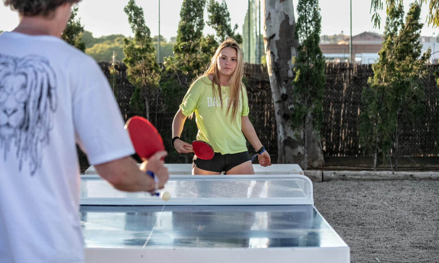 Two people playing table tennis outdoors with trees and a fence in the background at Gavina