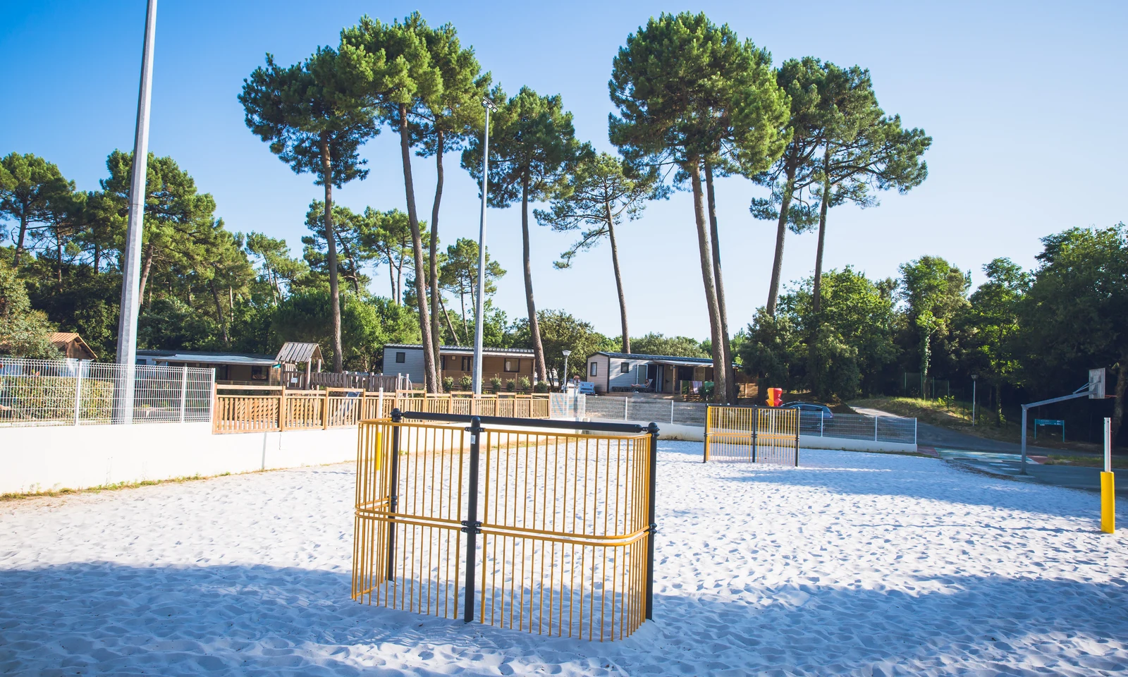 A sandy sports court with goalposts amidst tall trees, surrounded by wooden cabins at Domaine de Montcalm