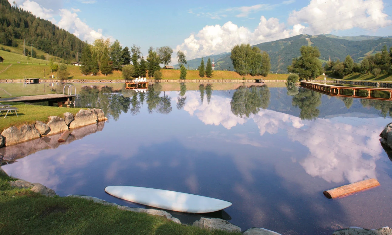 Calm lake reflecting trees, mountains, and clouds, with a wooden dock and lounge chairs on grassy shore at Sportcamp Woferlgut