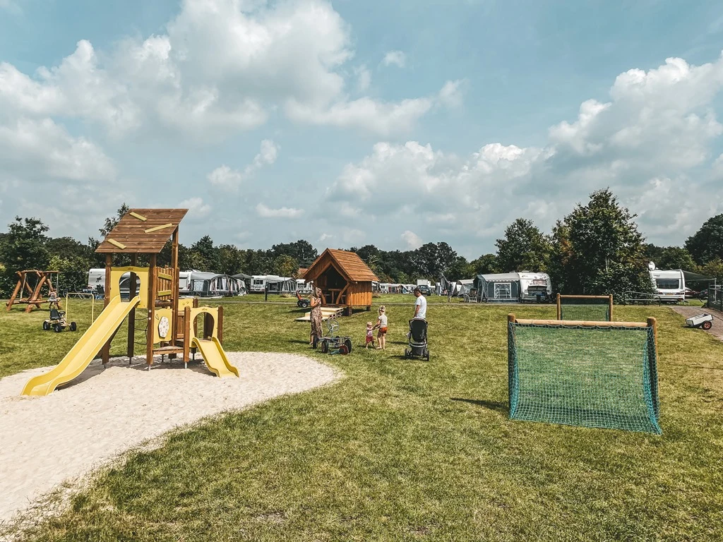 Playground set with slides and swings, children playing nearby, surrounded by grassy field with caravans and trees at Dal van de Mosbeek