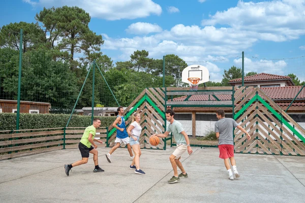 People playing basketball on an open outdoor court surrounded by trees and buildings at Au Lac de Biscarrosse