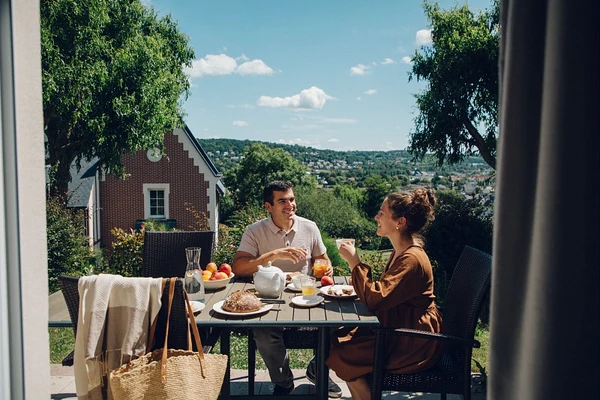 Two people dining outdoors at a table, chatting amidst green trees and a hillside view at Houlgate Premium Residence et Spa