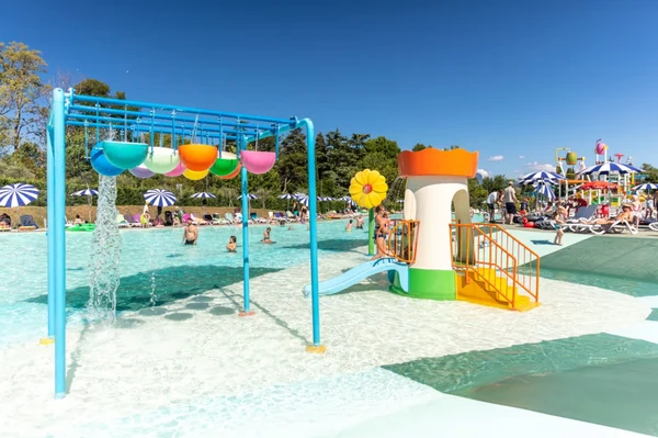 Children’s splash pad with colorful water buckets and slide; people relax under umbrellas in a sunny poolside area at Cisano/San Vito
