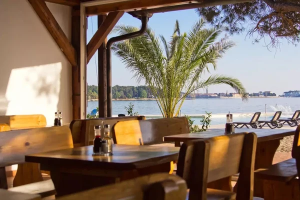 Wooden tables under a shaded patio facing a beachside with loungers and a palm tree at Boutique Camp Materada Beach