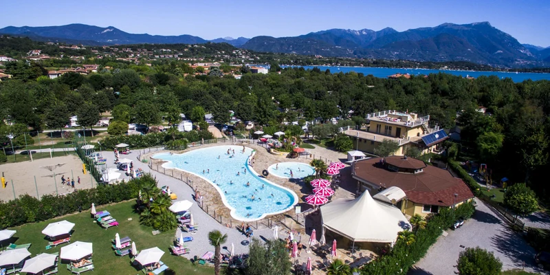 Swimming pool with sunbathers and swimmers, surrounded by loungers and umbrellas, located within a lush, mountainous lakeside campsite at Baia Verde