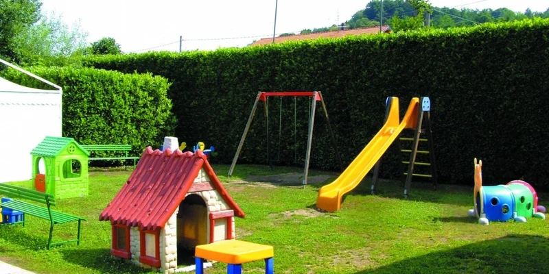 Playground equipment including a slide, swings, playhouses, and a tunnel caterpillar on grass with hedge backdrop at Okay Lido