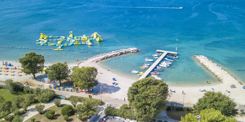 Inflatable water park on calm blue waters, surrounded by beach, trees, and anchored boats at Bi-Village