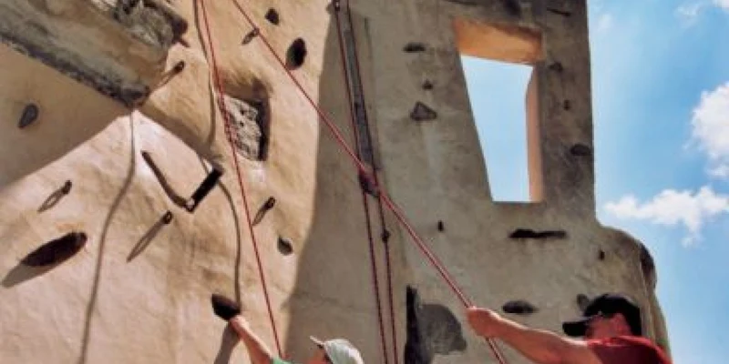 Two people rock-climbing on an artificial wall under a blue sky, with safety ropes secured at Airotel Pyrénées
