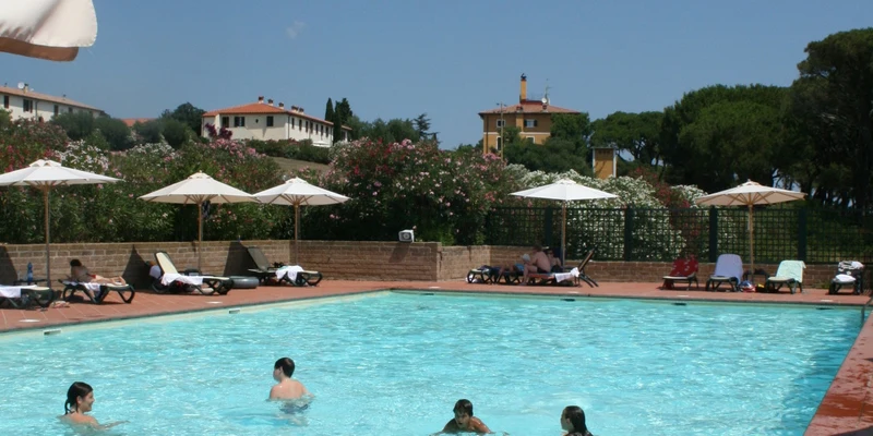 Several people swim in a pool surrounded by deck chairs and umbrellas, with buildings and trees in the background at Tenuta delle Ripalte