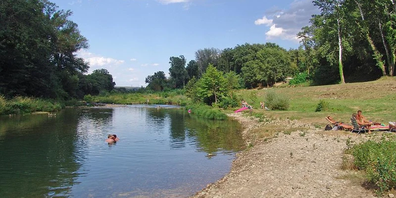 River flows gently, with people swimming and relaxing on the grassy and rocky bank, surrounded by lush trees at Slow Village Provence Occitane 