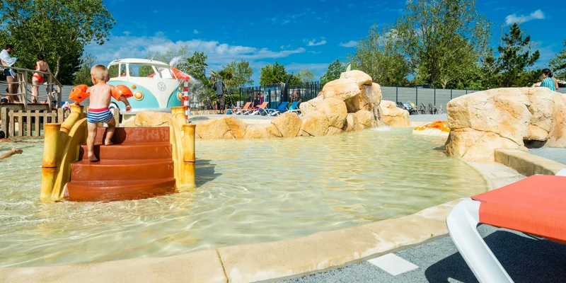 Child climbing a small water slide at a splash park surrounded by rocks and lounge chairs at Le Jard